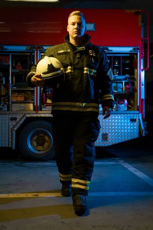 Full-length photo of young firefighter with helmet in his hand looking into camera while standing near fire truckの写真素材