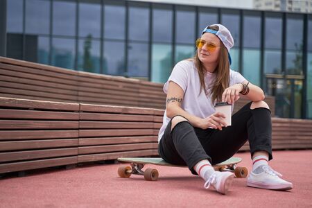 Photo of sports brunette in sunglasses in her hands sitting on skateboard on sports field near wooden bench on summer dayの写真素材