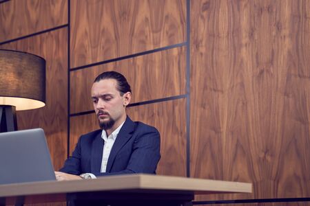 Photo of young businessman sitting at table with laptop on background of wooden wall.の写真素材