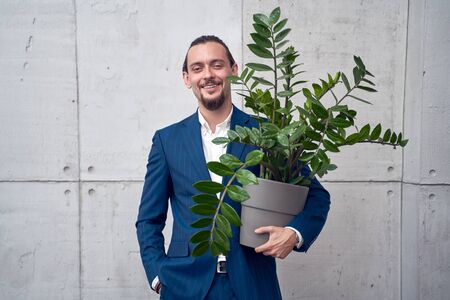 Image of happy brunet man in business suit with houseplant in hand on background of gray concrete...