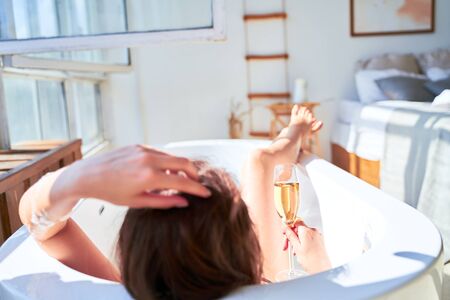 Image from back of woman with wine glass with wine in her hands lying in bath.