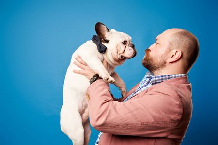 Bald man in jacket with french bulldog in his arms on empty blue backgroundの写真素材