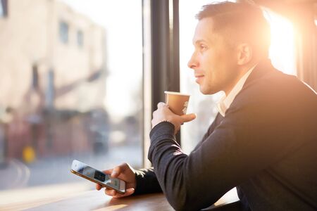 Man with phone and glass of coffee in his hands sitting near window in cafe during dayの写真素材