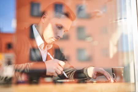 Male freelancer with phone and tablet sitting in cafe, building reflectionの写真素材