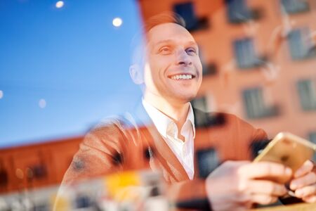 Happy male with phone in his hand sitting in cafe, building reflection in glass