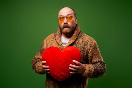 Male in pink glasses holds red toy heart while standing on empty green backgroundの写真素材