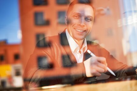 Young male businessman with phone and tablet sitting in cafe, building reflectionの写真素材