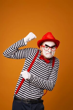 Sad mime with white face in red hat and striped t-shirt on blank orange backgroundの写真素材