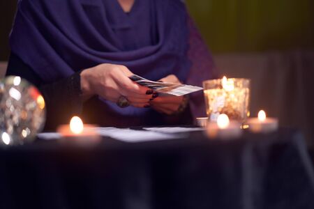 Close-up of fortune teller female divining on cards sitting at table with burning candles, magic...