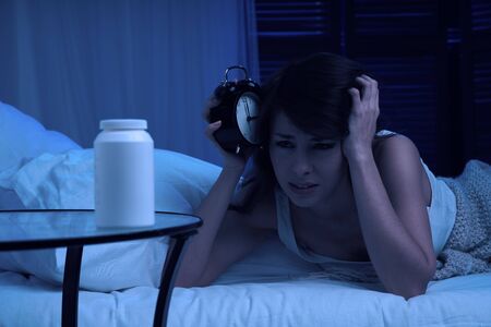 Unhappy brunette woman with insomnia lying on bed next to alarm clock and can of milk at nightの写真素材