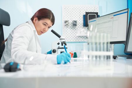 Young brunette lab technician looks at microscope in laboratory.