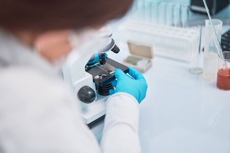 Close-up of female lab assistant in blue rubber gloves looking through microscope