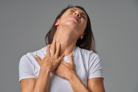 Woman touching chest and neck with eyes closed showing discomfort or sore throat indoorsの写真素材