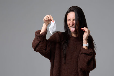 Female sticking out tongue with disgusted face holding used paper tissue on grey background