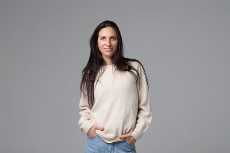 Portrait of smiling woman in white sweater standing with hands in pockets on grey background