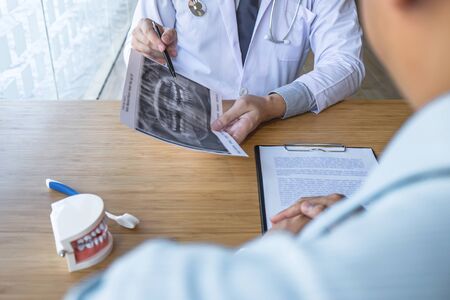 Professional Dentist showing  jaw and teeth the x-ray photograph and discussing during explaining the consultation treatment issues with patient and writing history list on report.の写真素材