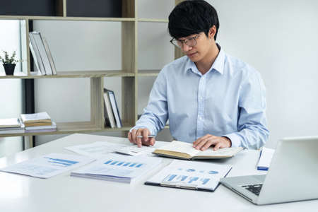 Young business man or accountant working on laptop with graph and report marketing research planning analyzing data statistics and calculator on office table.の写真素材