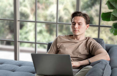 A good looking young man sits comfortably on a long sofa or laptop or notebook. A young man sits at work like working from home or accessing social media.の写真素材