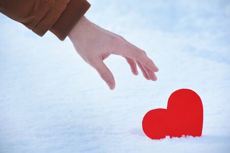 Lost red heart is alone on a valentines day, Heart on a cold winter snow, with a shining light around.Hand is picking up a red heart from the snow with care. Red heart stuck on a warm beautiful day.の写真素材