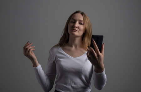 Beautiful young woman is making selfie photo with smartphone, golden haired woman over dark gray background, deep dark shadows, studio shotの写真素材