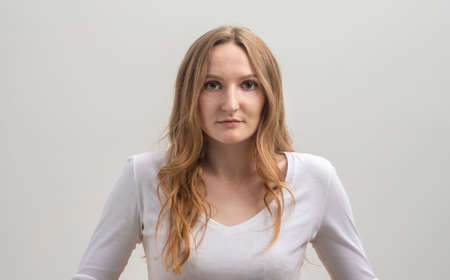 Studio portrait of serious focused caucasian young woman wearing white T-shirt, looking calmly and attentively at camera, over gray background, long golden hairの写真素材