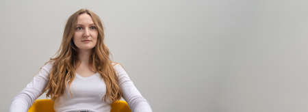 calm relaxed serious focused caucasian young woman wearing white T-shirt, looking calmly and attentively at camera, holding arms on chair over gray background, long golden hair, copy spaceの写真素材