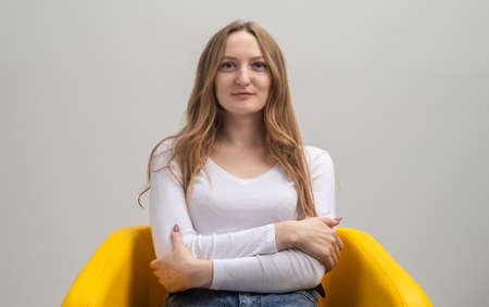 Portrait of a beautiful young woman with crossed arms thinking and looking at camera while sitting in yellow chair, gray background, studio shotの写真素材