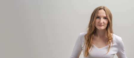 Studio portrait of serious focused caucasian young woman wearing white T-shirt, looking calmly and attentively at camera, over gray background, long golden hair, copy space, bannerの写真素材