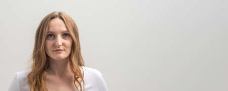 Studio portrait of serious focused caucasian young woman wearing white T-shirt, looking calmly and attentively at camera, over gray background, long golden hair, copy space, bannerの写真素材