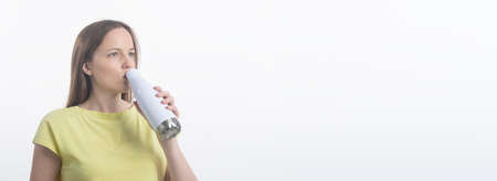 young caucasian woman drinking water from an eco white bottle in yellow t-shirt, white studio...