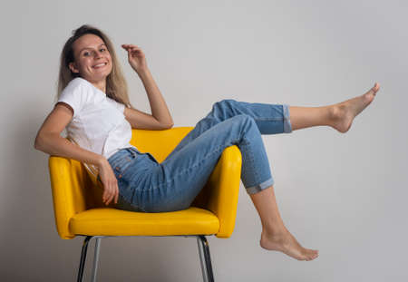 Beautiful young woman is sitting or lying on yellow chair with legs up and looking arrogantly at camera, studio shotの写真素材