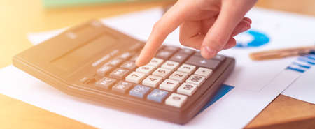 close up female accountant or banker hand making calculations sitting at office table with documents with tables on desk. Savings, finances and economy concept, counting on calculator, bannerの写真素材