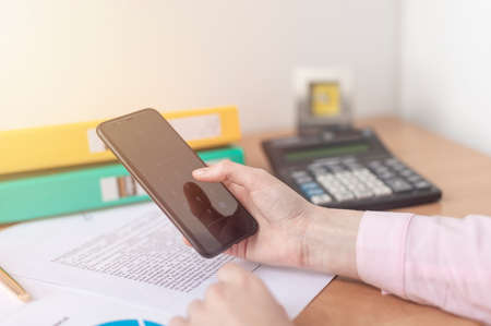 Close up female hands holding smart phone while connecting to wireless, businessman using technology sitting at wooden desk, people and modern devices everywhere, flare sun light, documents paperの写真素材