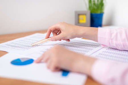 female accountant making calculations sitting at office table with documents with tables on desk pointing at digits on documents. Savings, finances and economy concept, working with documentsの写真素材