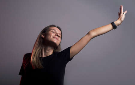 headshot of a young glad calm caucasian woman with closed eyes with satisfied expression waking up streaching her hand to the light. dark gray studio background, casula clothes.の写真素材