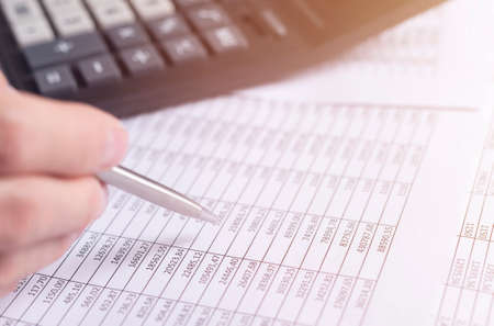 Accounting concept. male hand closeup with pen over financial documents with blurred calculator on...