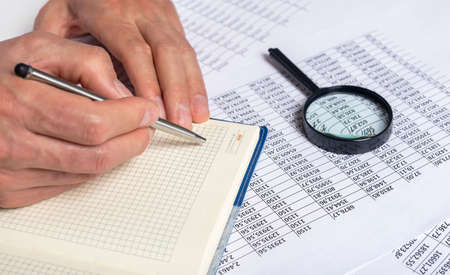 businessman writing and note on notebook with silver pen on table full of financial documents with digits and magnifying glass.の写真素材