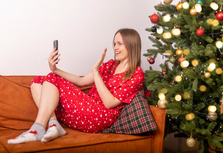 young beautiful woman waving hand and saying hello while chatting by video call on mobile phone, lying on sofa in pygamas, near decorated xmas tree. New year alone online at home.の写真素材