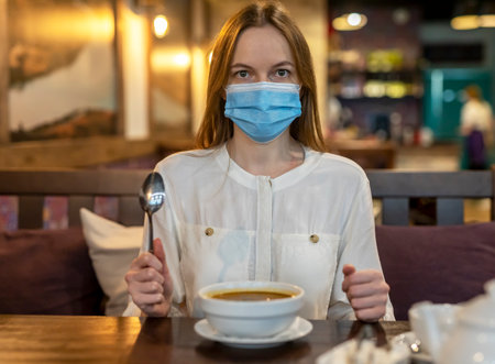 Caucasian woman in face protective mask at table in cafe eating soup. Public places during lockdown restrictionsの写真素材