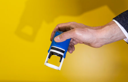 Male hand holding stamp for important document over yellow wall with shadow.