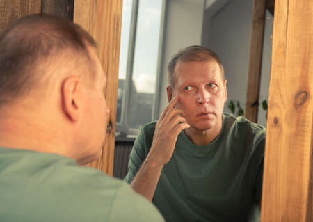 Mature middle-aged man looking in mirror reflection and watching wrinkles near eyesの写真素材