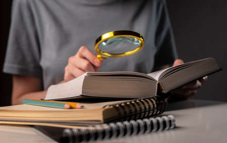 Female student hands close up, holding magnifying glass and book or textbook, searching for...