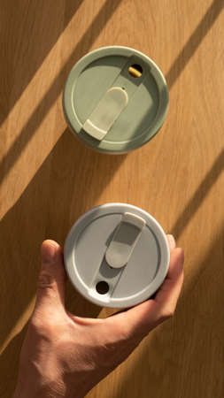 Male hand holding or taking eco coffe cup from wooden table with beautiful daylight from window, top...