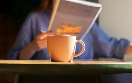 Coffee cup on wood table in cafe, blurred female hands with texbook or notebook, reading and writing, preparing for exam with notepad or planner closeup.の写真素材