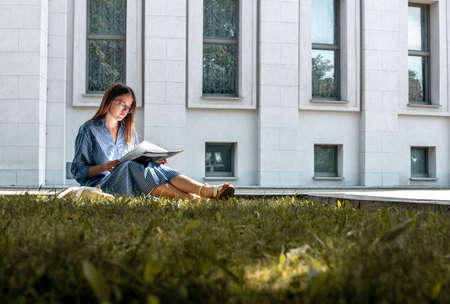 Young woman student sitting on grass near university building and learning, reading textbook. Young...