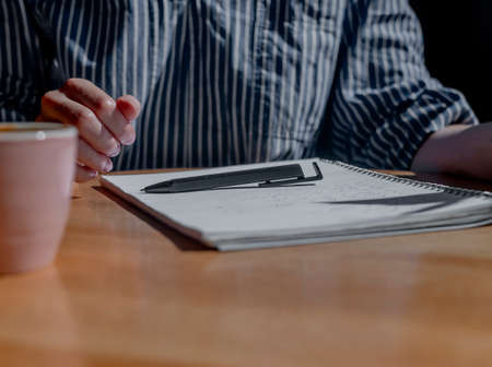 Planning concept. Woman hand with pen lying on notebook or planner on wooden desk with sunlight and coffee cup, close up.の写真素材