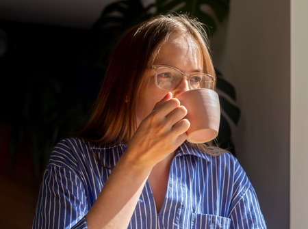 Woman in glasses sitting by wood table in coffee shop, holding cup and drinking. Sad tired female in...