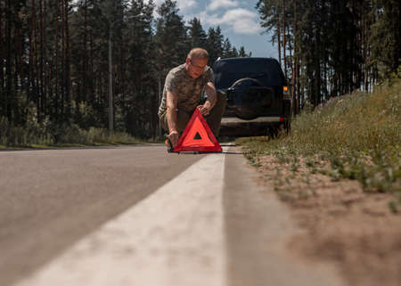 Driver man putting red triangle caution and warning sign on road near broken car.