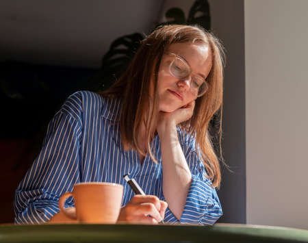 Smiling dreaming woman writing plans in notebook, sitting at wood desk in cafe with coffee cup and...