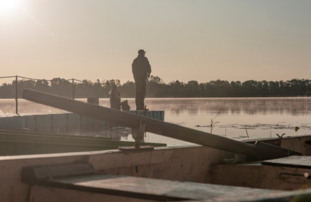 Fisherman fishing in foggy morning. Man silhouette resting with rod on river or lake in mist.の写真素材